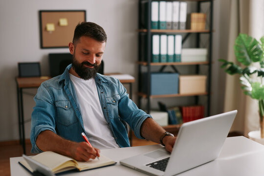 Smiling business professional writing in notebook and using laptop during productive work session at home