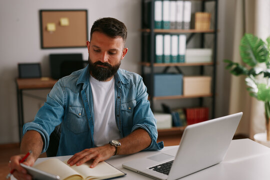 Concentrated professional writing in notebook and reviewing information on laptop in a modern home workspace
