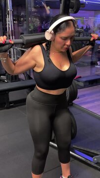 A young Latina woman performing barbell squats in a well-equipped gym. The footage captures her proper form, controlled movement, and focused expression as she works through the exercise. 