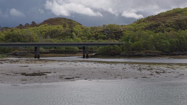 Telephoto static drone shot of the Morar Bridge spanning the tidal river, framed by lush green hillsides and a moody Scottish sky.