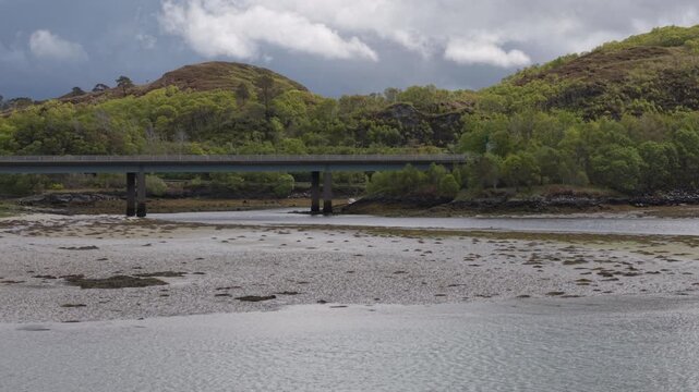 Telephoto drone flight pushing slowly over the Morar River toward the concrete span of the Morar Bridge, backdropped by lush, rolling Scottish Highland hills.