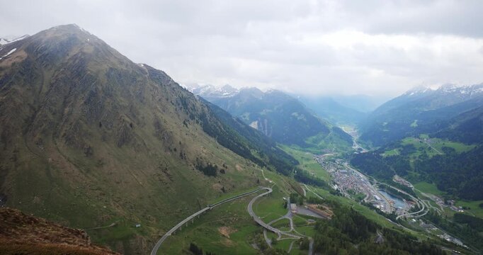 Gotthard Pass, Switzerland, May 18, 2026. Wide panoramic panning shot overlooking the Leventina Valley and the town of Airolo, showcasing mountain roads, green hillsides, and snowy peaks