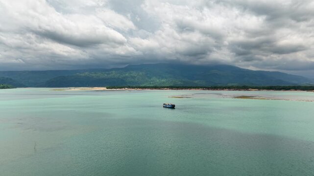 Aerial view of a lone boat navigating the turquoise waters of Tanguar Haor with lush green hills under a cloudy sky in Sunamganj District, Sylhet Division, Bangladesh.