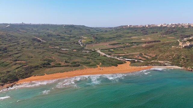 Ramla Beach in Gozo, Malta, features distinctive red sand and turquoise Mediterranean waters bordered by cliffs and hills, seen from a bird's-eye perspective on a sunny spring day.