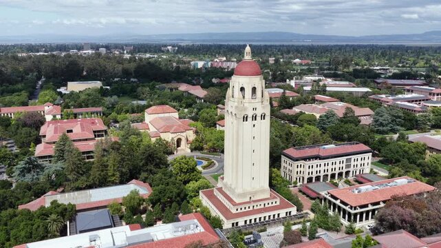 Hoover Tower with red dome rising above red tile campus buildings, lush tree canopy and Silicon Valley sprawl stretching toward distant mountains under cloudy sky. California. Aerial view. Quiet scene
