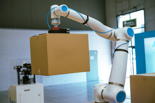 A white and blue industrial robotic arm lifts a cardboard box using a suction cup end effector during a logistics automation demonstration.