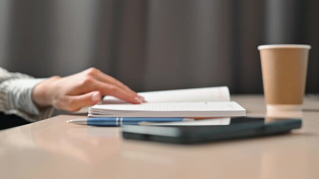A person is reading and taking notes at a desk. The workspace features notebooks, a coffee cup, and a smartphone, reflecting casual study habits