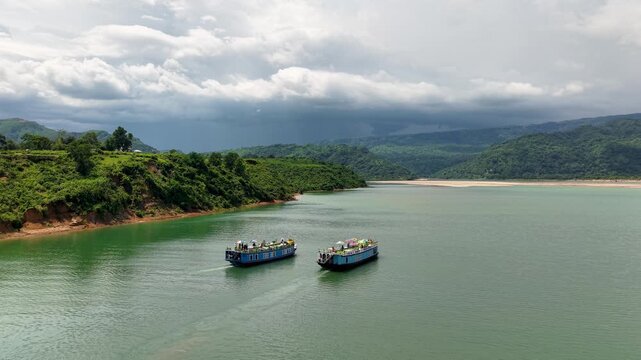 Aerial view of two traditional boats cruising on the calm waters of Tanguar Haor surrounded by lush green hills under a cloudy sky Sunamganj District, Sylhet Division, Bangladesh.