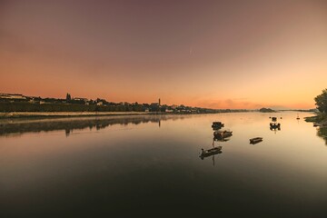 Fototapeta premium As the sun begins to set, serene waters mirror the sky's vibrant hues. Small boats drift gracefully on the river, with hills framing a picturesque scene of peace and beauty
