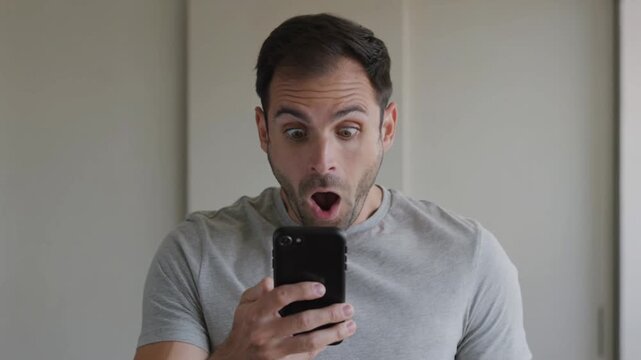 Surprised man expressing shock while looking at his smartphone screen indoors against a neutral wall