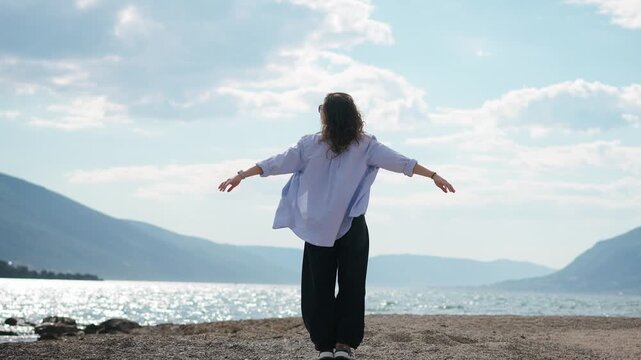 Slow motion shot of a young woman standing on the seashore with arms outstretched, while her hair and shirt flow in the wind, expressing freedom and connection with nature