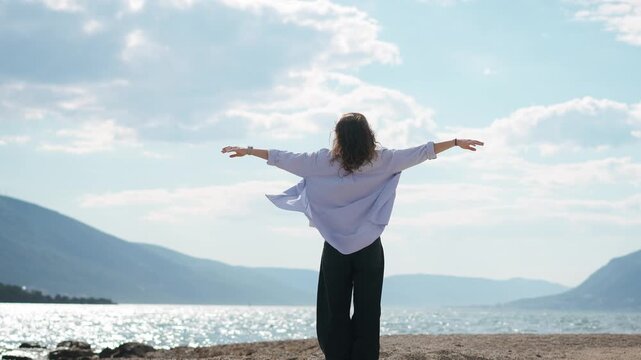 Slow motion shot of a young woman standing on the seashore with arms outstretched, while her hair and shirt flow in the wind, expressing freedom and connection with nature