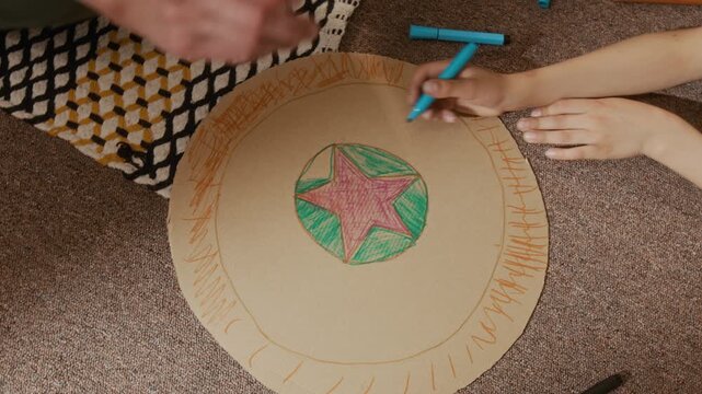 Top down shot of child and parent drawing star on cardboard circle together using colorful markers. Video is perfect for family bonding campaigns, creative education promos and parenting workshops