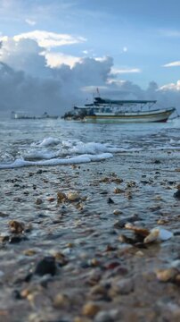 Clear ocean wave rolling on sand shell shore close-up. Defocused tourist boat background on tropical beach destination. Tanjung Benoa, Bali, Indonesia