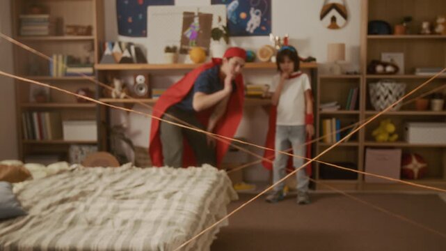 Zoom out shot of young man and boy wearing superhero capes planning how to cross yarn laser maze in bedroom. Footage ideal for parenting, family bonding, and creative play campaigns