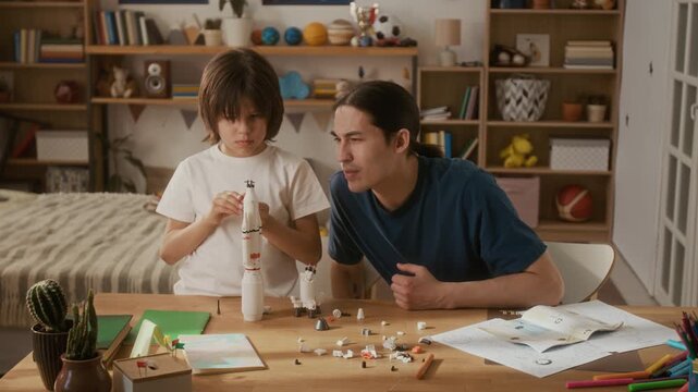 Young adult man and multiethnic boy assembling toy space rocket model at wooden table.Clip represents STEM education and family bonding for toy marketing or parenting campaigns