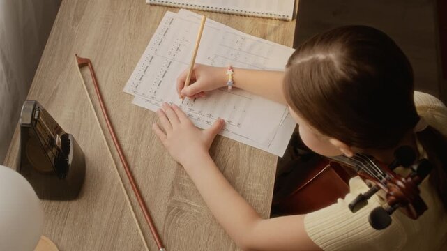 High angle view of Caucasian child writing notes on sheet music with pencil at desk during cello practice. Footage is ideal for promoting music schools, elementary education, and private tutoring