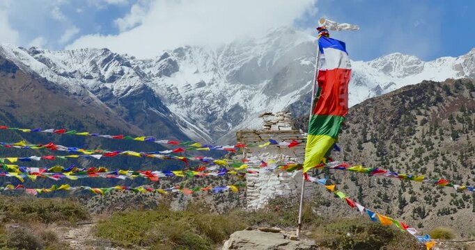 Sideways drone shot capturing a traditional Buddhist chorten with vibrant Lung ta and Darchog flags in Mustang, Nepal, as it is fluttering intensely against a cold, misty Himalayan mountain