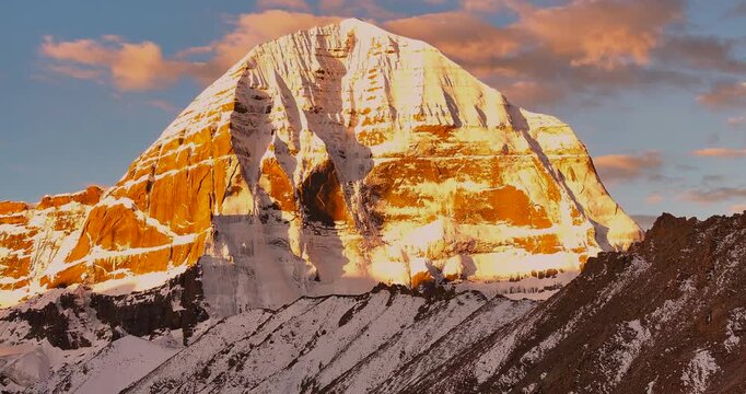 A slow lateral drone shot captures Mount Kailash in Tibet filmed against direct sunlight, highlighting warm yellow and brown tones across the snow-covered surface and rocky peak under a clear sky.