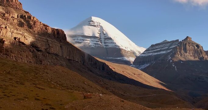 A drone push-in shot captures the iconic, snow-covered peak of Mount Kailash rising beyond red sandy hills and rugged desert terrain in Tibet under clear daylight conditions.