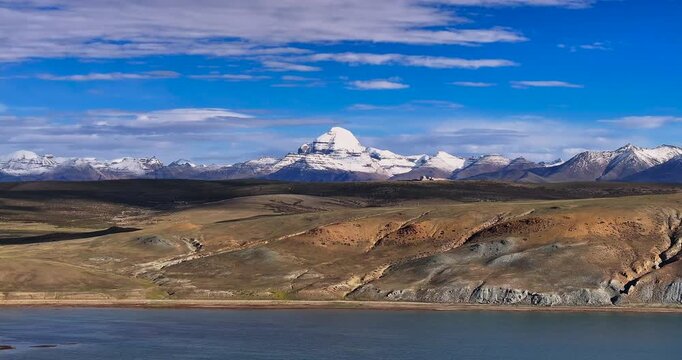 A lateral drone glide shot captures the vibrant blue water of Lake Manasarovar alongside the Mount Kailash mountain range in Tibet, revealing arid terrain, rolling hills, and snow-covered peaks.