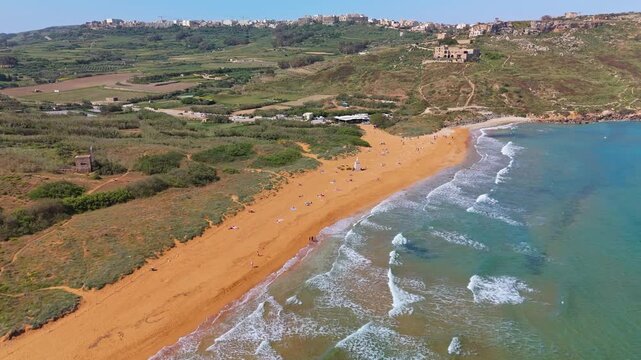Ramla Beach on Gozo features its distinctive reddish sand and clear turquoise waters along the Mediterranean coastline, with gentle waves breaking near the shore in this drone view.