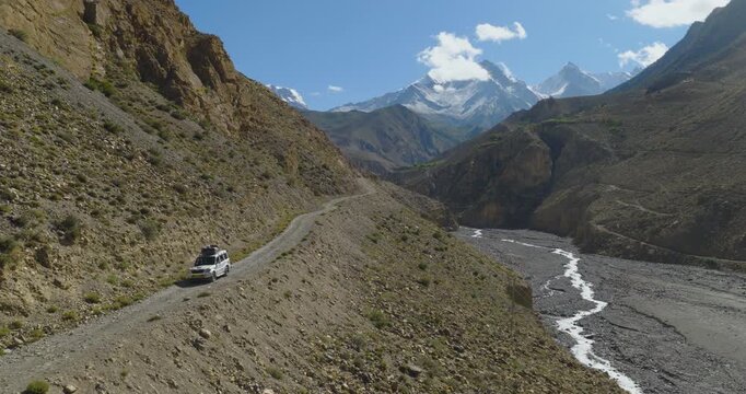 Aerial drone pull-out shot capturing an SUV driving on a winding road through the rugged terrain of Mustang, Nepal, revealing rolling hills, a flowing river, and majestic Himalayan mountains.