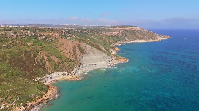 Coastline near Ramla Beach in Gozo, Malta, features turquoise Mediterranean waters bordered by rocky cliffs and green hills, as seen from a drone on a sunny day.