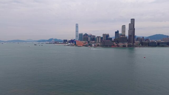Aerial view across Victoria Harbour toward Tsim Sha Tsui skyline with ICC tower Kowloon waterfront and boats below distant hills in Hong Kong