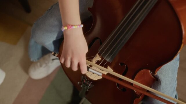 View from above of child girl playing cello during music lesson at home, focusing on finger placement and bow movement. Footage ideal for music education, online tutoring, and arts school promotions