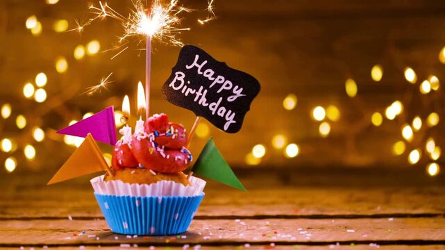 Festive birthday cupcake with pink frosting, burning candles, and sparkling sparkler stick close up on wooden table against warm golden bokeh background, celebratory holiday greeting card concept 4K 