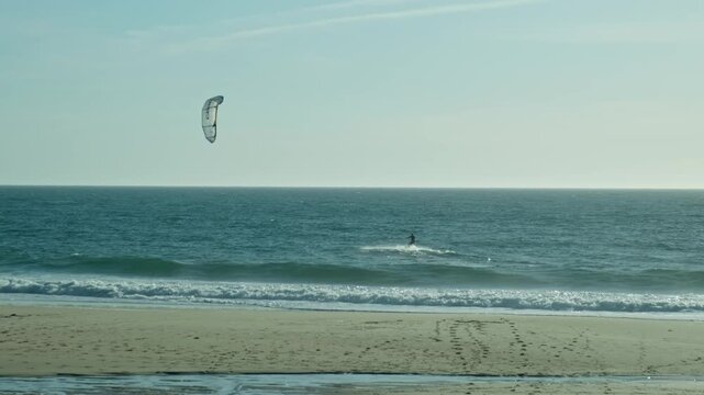 Sportsman go kitesurfing in Atlantic Ocean, slow motion. Extreme sports, skateboarding on vacation on sandy beach in windy weather. Extreme Kitesurfing. Man Kite Surfing Against blue Sky.