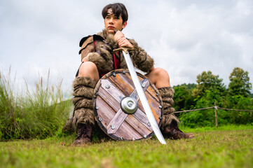 Obraz premium Male ancient warrior in full armor holding sword and shield sitting in woodland 