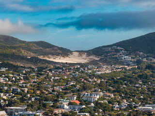 Fototapeta premium Soft daylight shows houses, villas, and roads near Hout Bay or Noordhoek, Cape Town. A pale sand dune cuts the saddle between rugged hills, with patchy clouds above.