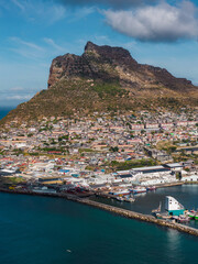 Obraz premium Aerial scene of Hout Bay Harbour near Cape Town, South Africa, with the Sentinel peak, hillside houses, colorful boats, and warehouses under bright midday light and clouds.