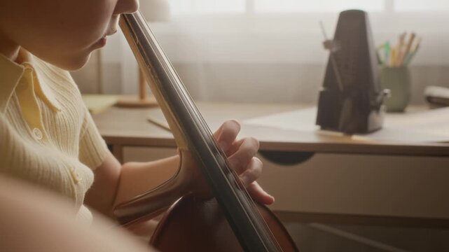 Close up of Caucasian child practicing cello at home desk with ticking metronome in background. Footage is ideal for music school commercials, online education platforms, and childhood development ads