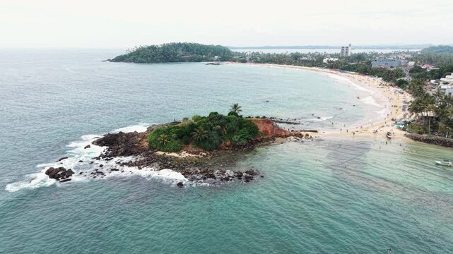Aerial view of Parrot Rock, a rocky island connected to the sandy beach by a narrow sandbar with people, turquoise waters and crashing waves Mirissa, Southern Province, Sri Lanka.