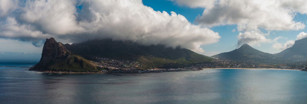 Aerial view of Hout Bay, the Sentinel on the left, and Little Lion's Head on the right in Cape Town, South Africa. Soft daylight casts chiaroscuro over calm Atlantic water.