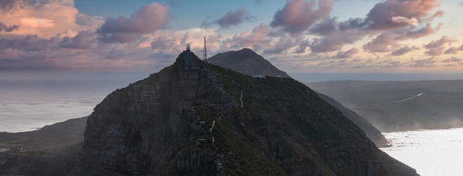 Aerial view shows Cape Point in Cape Town, South Africa, with cliff top lighthouse, winding footpath, steep drop offs, soft sunset light, layered clouds, and misty sea.