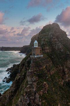 Aerial view of Cape Point Lighthouse on a rugged cliff near Cape Town, waves churn below and pink blue sunset clouds hang above, soft fading daylight sets a moody scene.