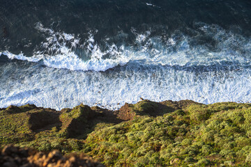 High angle shot looking down at the blue ocean waves breaking into white foam against a rocky cliff covered in green plants and shrubs during daylight. © JULIA