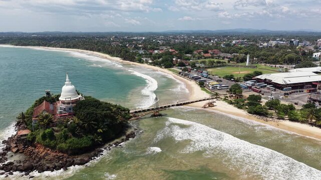 Aerial view of Parewi Duwa Temple on a rocky island connected by a bridge to the sandy beach and coastal city Matara, Sri Lanka.