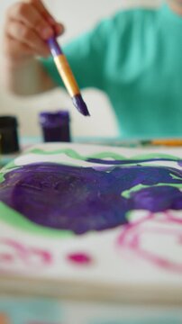 Vertical close up of a child's hand with a paintbrush painting. 9:16 video of a kid painting a colorful picture, developing creativity in kindergarten. Little kid drawing with paint and a small brush.