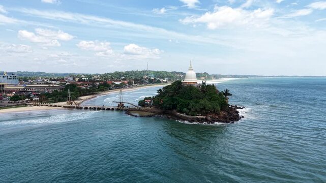 Aerial view of Parewi Duwa Temple on a small island connected by a bridge to the beach with a white stupa and lush greenery in Matara, Sri Lanka.