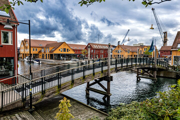 Fototapeta premium Scenic waterfront canal with colorful historic buildings and moored boats under dramatic sky, Kristiansand, Norway