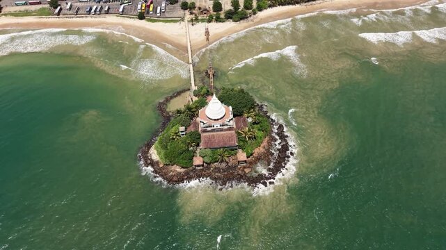 Aerial view of Parewi Duwa Temple on a small island connected by a bridge to the sandy beach and turquoise ocean in Matara, Sri Lanka.