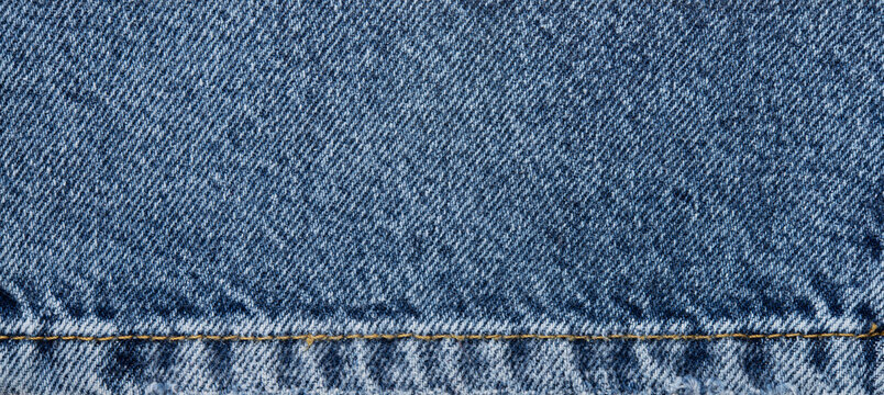 Blue denim fabric texture with seam, classic jeans cloth background with yellow stitching and frayed edge.