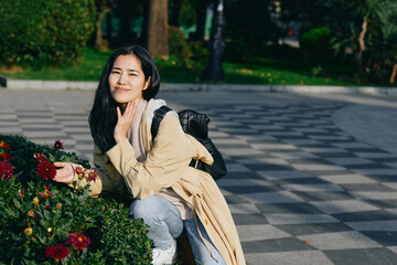 Fototapeta premium Woman crouching beside a flower bed in a park, wearing a light coat and backpack, enjoying a sunny day among blooming plants and greenery, relaxed pose and gentle smile