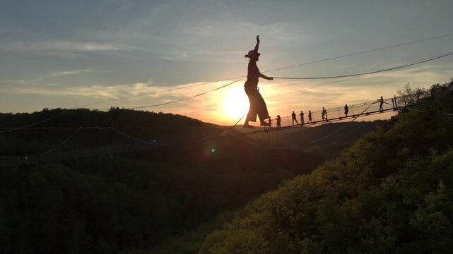 Highline Aerial Shot. Brave Girl Tightrope Walker is on a rope stretched between Mountains at high altitude, Sunset Time