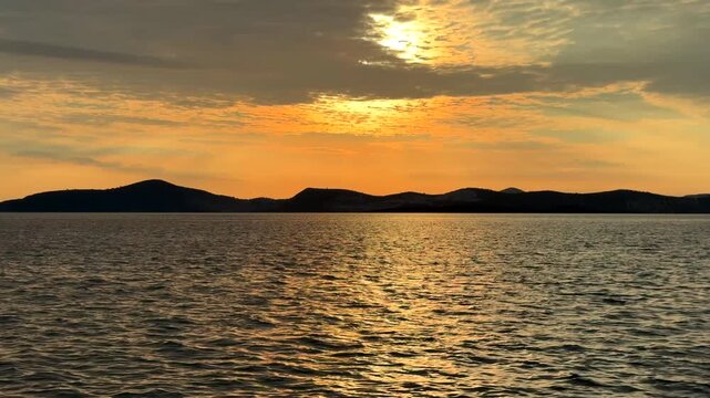 A stunning golden sunset over the Aegean Sea in Gulluk, Turkey. Bright sunlight breaks through scattered clouds, casting a warm orange glow over the rippling water and dark mountain silhouettes